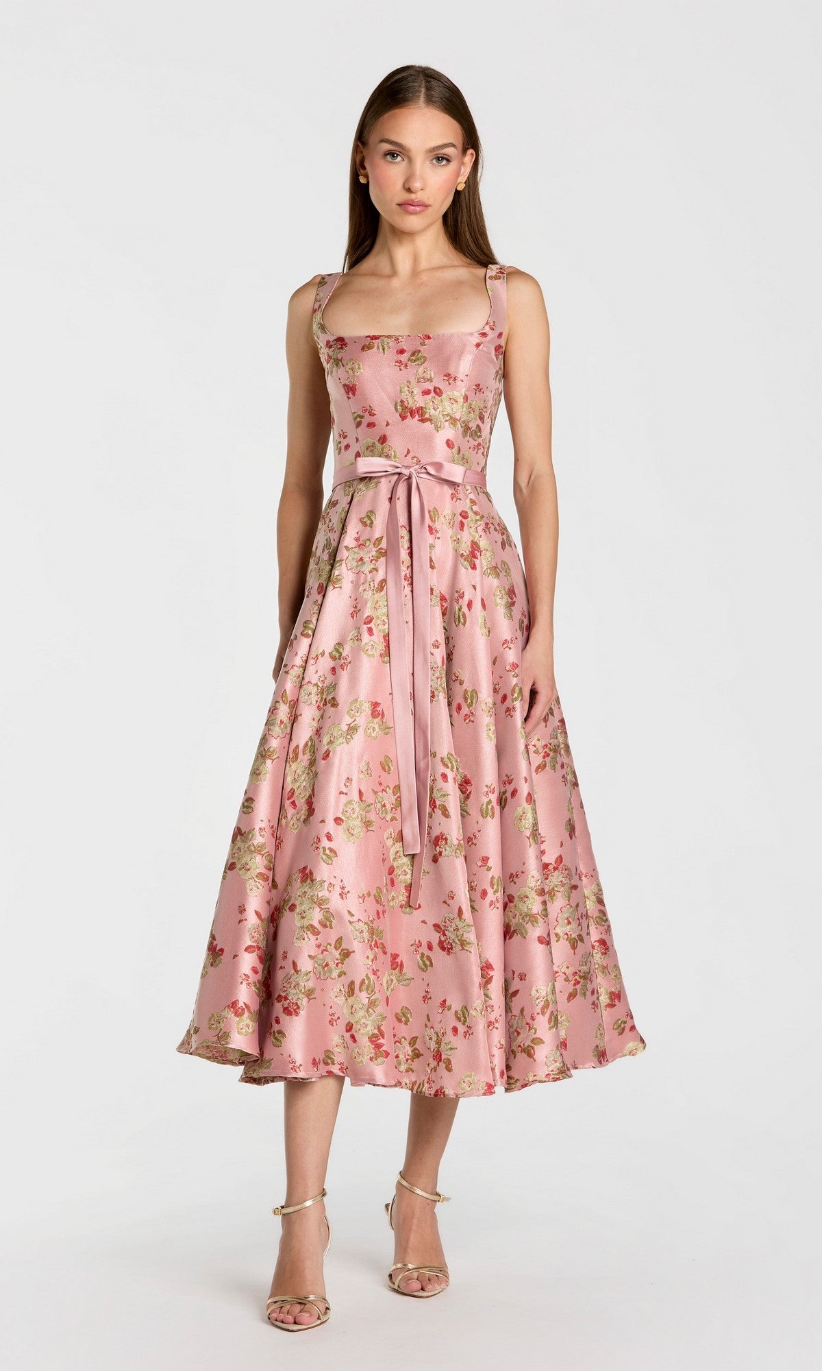 A woman stands against a plain white background wearing the Midi Party Dress: Mac Duggal 27767 in Pink Multiโa sleeveless floral midi dress featuring a fitted bodice, bow tie at the waist, and paired with strappy heels.